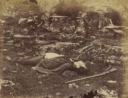 Photographie en noir et blanc d'un soldat mort, allongé sur le dos, sur un terrain plan.