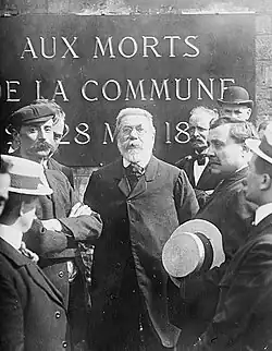 Photographie d'un homme entouré par d'autres hommes devant une plaque commémorative.
