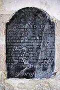Photographie en couleurs d'une plaque en ardoise scellée dans un mur et couverte d'inscriptions.
