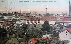 Ancienne carte postale colorisée. Vue en plongée sur des usines desquelles dépassent cinq longues cheminées