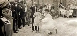 Photographie noir et blanc. Dans un cadre champêtre d'un homme genou à terre devant une fillette recevant une couronne en présence d'une assemblée.