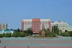 Photographie d'une grande place avec au centre-droit une statut de Lénine et derrière un grand bâtiment brutalistes rouge et blanc d'une dizaine d'étages, sur fond de ciel dégagé.