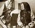 Photographie sépia d'une quadragénaire vêtue d'un costume traditionnel, comportant notamment une coiffe de tissu complexe, assise derrière une vieille femme portant un fichu et échangeant avec elle un regard complice.