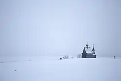 Photographie d'un paysage plat entièrement blanc, avec seulement une église en bois qui sort de ce paysage enneigé.