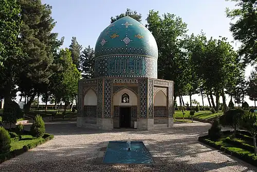 Un bâtiment élégant de style islamique, avec une moitié inférieure composée de trois porches à arcs courbés, et un dôme bleu vif au sommet, se dressant sur une allée au milieu d'un jardin verdoyant.