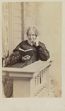  Photographie sépia d'une femme blanche d'âge mûr portant un bonnet blanc, assise et lisant sur un balcon