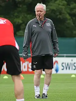 Homme, debout, les cheveux blancs, sur un terrain de football, en habit sportif, un sifflet à la bouche.