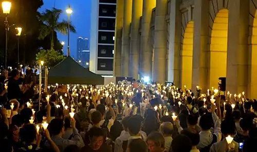 Quelques centaines des mille cinq cent manifestants chinois présents pour le septième jour du décès de Li Wangyang, massés au premier plan à droite devant les arcades éclairées du Conseil législatif à Hong Kong, et tenant des cônes de papier enserrant une bougie. Buildings, palmiers, lampadaires et ciel bleu-nuit éclairée en arrière-plan.