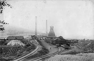 Photo noir et blanc de petits bâtiments, avec deux cheminées et tour pyramidale en bois fermée..