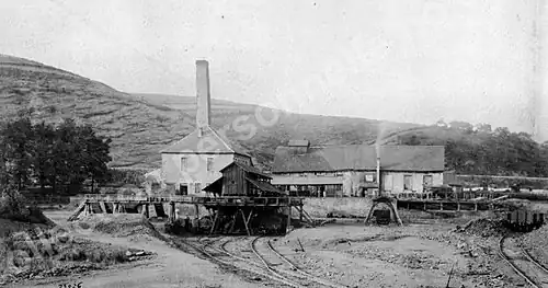 Gravure en noir et blanc : un bâtiments avec toit à quatre pans est doté d'une cheminée de base carré à côté d'un autre bâtiment tout en longueur muni d'un clocheton, des voies ferrées passent devant les bâtiments.