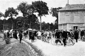 Photographie en noir et blanc montrant un attroupement sur une route devant une auberge.