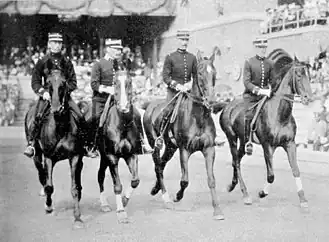 Photo noir et blanc. Devant une tribune, quatre cavaliers avancent alignés de front, vus de face. Tous sont des militaires.