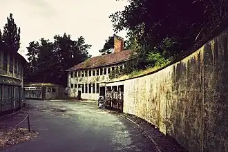 Un bâtiment en arrière-plan, à deux étages et toit en tuiles. À l'avant-plan, un mur incurvé suit la route.