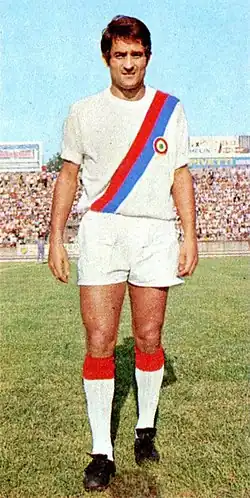 Un joueur qui marche sur le terrain en regardant l'objectif, portant un maillot blanc avec rayure diagonale bleue et rouge.