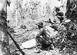 Photo noir et blanc d'un militaire couché avec un fusil mitrailleur. Deux autres militaires sont accroupis à ses côtés.