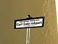 Photographie d'un panneau signalétique indiquant en hongrois, en lettres noires sur fond bland "XIII. kerület, Újlipótváros" et "Carl Lutz rakpart".