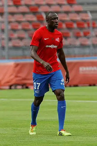 Photographie d'un homme sur un terrain de football avec un maillot rouge, un short bleu, des chaussettes bleues et des chaussures de football jaunes. En arrière-plan, une tribune