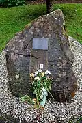 Photo rapprochée d'une large pierre comportant en son centre une plaque commémorative grise. La pierre est entourée de petits graviers blancs et des fleurs jaunes et roses sont déposées à sa base.