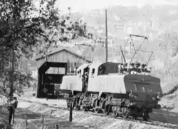 Photographie en noir et blanc de la locomotive vue de trois quarts en train de sortir de sa remise en bois.