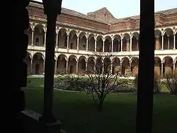 Image photographique d'un cloître. Deux colonnes au premier plan encadrent une cour verdoyante et un rond central avec au fond deux ailes à angle droit d'un édifice constitué s'arcades superposées sur deux niveaux surmontés par une toiture.