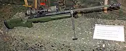 Photographie couleur. Exhibition d'un fusil de précision monté sur bipied accompagné, sur le même plan, d'une légende indiquant en plusieurs langues (allemand, anglais, français et italien) le calibre de l’arme. Le canon de l'arme est pointé vers la droite. L’ensemble repose sur un filet de camouflage