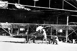 Photographie en noir et blanc d'un biplan monomoteur et de deux hommes devant un grand hangar ouvert