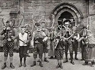 Photo en noir et blanc d'une douzaine d'hommes qui posent debout devant l'objectif, plusieurs sont vêtus de pantalons bariolés et portent dans les mains des bois de renne