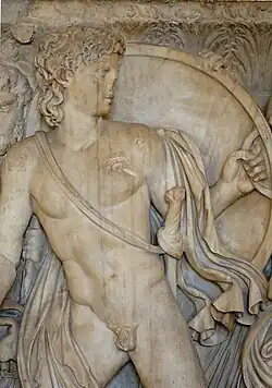 Photographie d'un détail d'un bas-relief en pierre. Achille est représenté comme un jeune homme imberbe aux cheveux ondulés et mi-longs. Il est debout, tourné vers la droite. Il est nu, à l'exception d'un tissu drapé qui passe sur son épaule gauche et dissimule en partie son bras gauche, mais passe dans son dos en laissant voir son torse et son sexe, avant de revenir par-dessus sa cuisse droite. Achille porte une épée rangée dans un fourreau accroché en bandoulière autour de son épaule gauche. Achille regarde vers la droite et lève la main gauche avec laquelle il tient un grand bouclier rond. Le bas des jambes d'Achille et son bras droit sont hors du cadre de la photo. Derrière Achille, on entrevoit en haut des chapiteaux de colonnes corinthiennes à motifs floraux, et, en bas, des draperies.