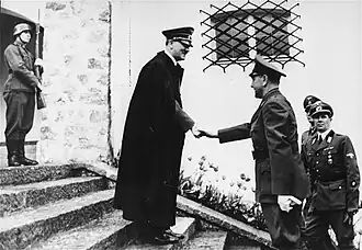Photo noir et blanc, prise le 9 juin 1941 à Berchtesgaden, en Allemagne. Au centre de la photo, Adolf Hitler, portant un long manteau noir et une casquette militaire, serre la main d'Ante Pavelić (à droite), dirigeant de l'État indépendant de Croatie. Les deux hommes sont de profil, debout, sur les marches de l'escalier menant à l'entrée de la résidence secondaire de Hitler. Un soldat en arme se tient debout, en haut de l'escalier (en haut à gauche de la photo). Au second plan, deux officiers allemands (en bas à droite de la photo) assistent à la scène. L'arrière-plan de la photo est constitué d'un mur blanc percé d'une fenêtre grillagée (au-dessus de la tête du dirigeant croate).