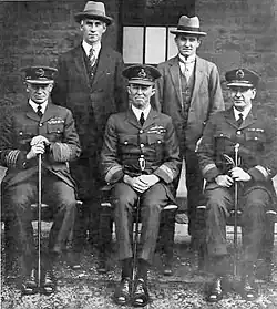 Portrait formel de cinq hommes, trois assis et portant des uniformes militaires avec des casquettes à visière, deux debout et portant des vêtements et des chapeaux civils.
