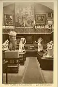Carte postale en sépia et blanc montrant l'intérieur d'un musée, statues sur des socles et tableaux aux murs.