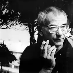 Un homme de face fumant une cigarette et regardant à sa gauche. Il porte de grande lunettes et a peu de cheveux.