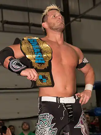 Un catcheur brun aux cheveux teints en blonds torse nu avec une ceinture de champion sur l'épaule droite.