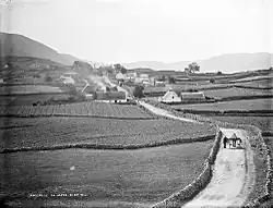 Photographie en noir et blanc d'un paysage de campagne, à gauche et à droite des champs de culture séparés par des murets de pierre, entre les deux, une route où circule une charrette, au fond des habitations