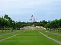 Photographie en perspective du parc, qui s'élève par de larges escaliers se terminant par une cour et un cercle de cinq drapeaux.