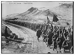 Soldats américains marchant sur un chemin de gravier près de Vladivostok, dans un paysage entouré de monts, avec un soldat tenant le drapeau américain.