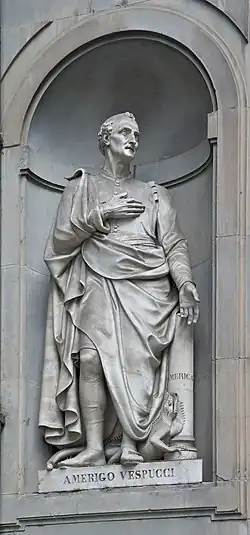 Photographie d'une statue en pied d'un homme assez âgé la main droite sur la poitrine. À ses pieds se trouve une sorte d'iguane.