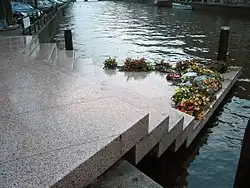 Photo en couleurs (teinte bleutée dominante), prise à Amsterdam en novembre 2003, et montrant le « Homomonument » : un monument dédié aux victimes homosexuelles du nazisme. La photo en montre seulement une partie : un assemblage de cinq dalles en granit rose qui forment un escalier triangulaire permettant d'accéder, du trottoir, au bord d'un canal (de gauche à droite sur la photo). La dernière marche, une plate-forme triangulaire, qui semble flotter sur l'eau du canal, est en partie recouverte de gerbes de fleurs. L'arrière-plan de la photo est constitué par l'étendue d'eau du canal.