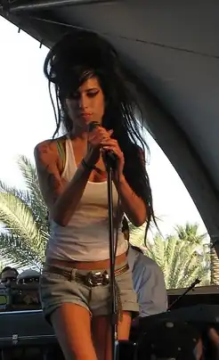 Chanteuse en position debout, tenant un micro sur pied dans ses mains, regardant au sol, image en couleur.