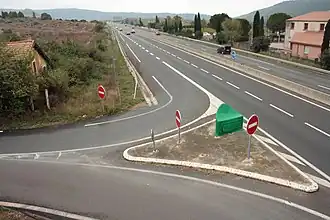 Vue diurne, en plongée (depuis un pont voisin), de l'ancien bâtiment voyageurs de la gare Salelles-du-Bosc (sur la gauche), jouxté par la bretelle de sortie à angle droit de l'autoroute.