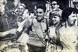 Photo en noir et blanc d'un homme en tenue de cycliste avec une femme blonde à sa gauche. D'autres personnes sont en arrière-plan de la photo.