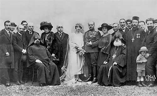 Photographie en noir et blanc d'une vingtaine de personnes entourant un couple marié au centre. La mariée porte une robe blanche, un voile et un bouquet de fleurs dans la main, tandis que le marié porte un costume militaire.