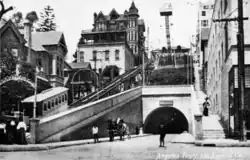 Photographie en noir et blanc. On voit quelques passants ainsi qu'une calèche.