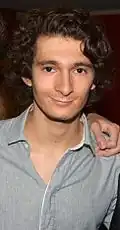 Portrait photographique en buste d'un homme d'une vingtaine d'années, fixant le photographe avec un léger sourire. Il porte une chemise gris claire.