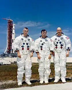 Trois hommes debouts en combinaison spatiale. Fusée sur site de lancement à l'arrière plan.