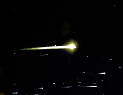 Des bandes blanches de lumière, avec des points lumineux sur le côté droit, remplissent le bas du cadre. Une grande sphère jaune striée se trouve au centre du cadre. L'arrière-plan est un espace noir.