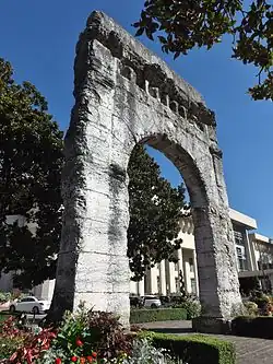 Photographie en couleurs des vestiges d'un arc de triomphe romain ; gra, d bâtiment à l'arrière-plan.