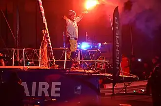 Dans la nuit, entouré d'embarcations, debout à l'avant de son bateau, il brandit un feu à main rouge.