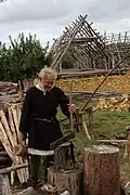 Photographie montrant un homme costumé, équipé d'un maillet et d'un départoir, se préparant à fendre une bûche, avec un bâtiment en pierres en construction, surmonté d'une charpente formée de troncs.