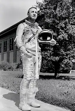 Un homme debout, en combinaison spatiale, son casque sous le bras droit.